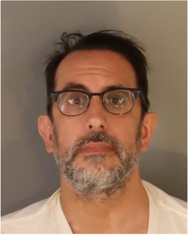 Booking photo of male, white t shirt, dark hair, black round glasses, and salt and pepper beard/mustache 