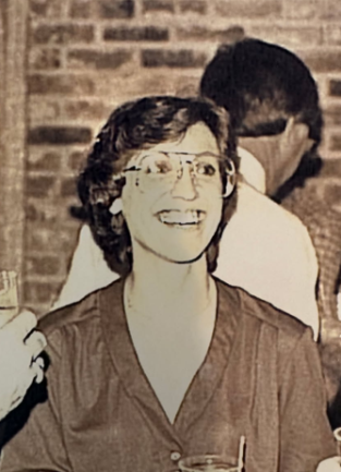 Woman smiling looking off to her left with 70s/80s style glasses on. The photo is sepia toned.